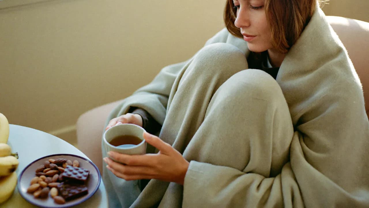 Une femme blottie sous un plaid avec une tisane, du chocolat noir et des amandes à portée de main