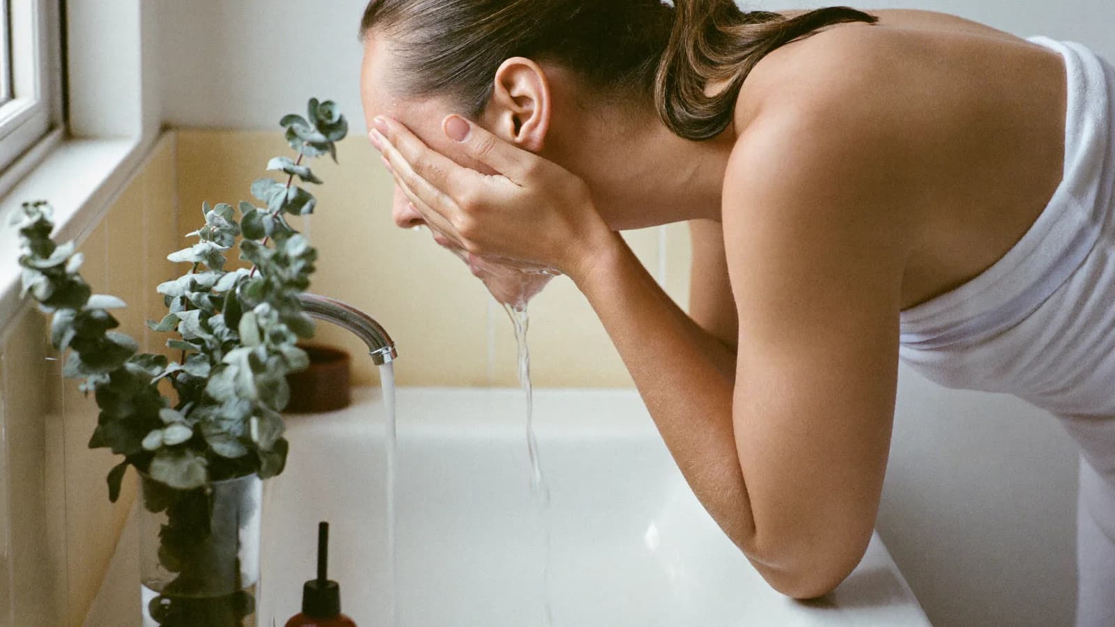 Une femme se passe de l'eau sur le visage au-dessus d'un lavabo, une serviette sur l'épaule
