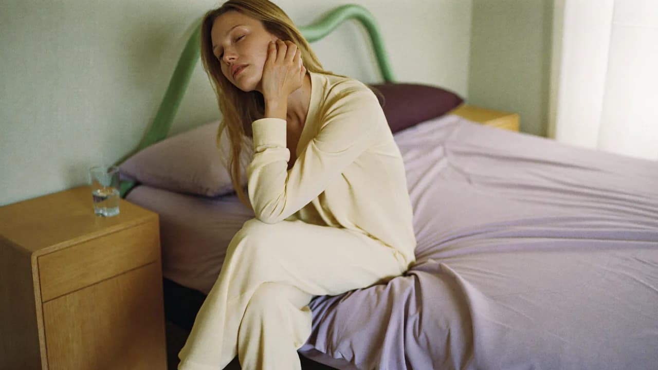 Une femme assise au bord de son lit au réveil, les yeux mi-clos, une main contre le cou, un verre d'eau sur la table de nuit, dans la lumière pâle du matin