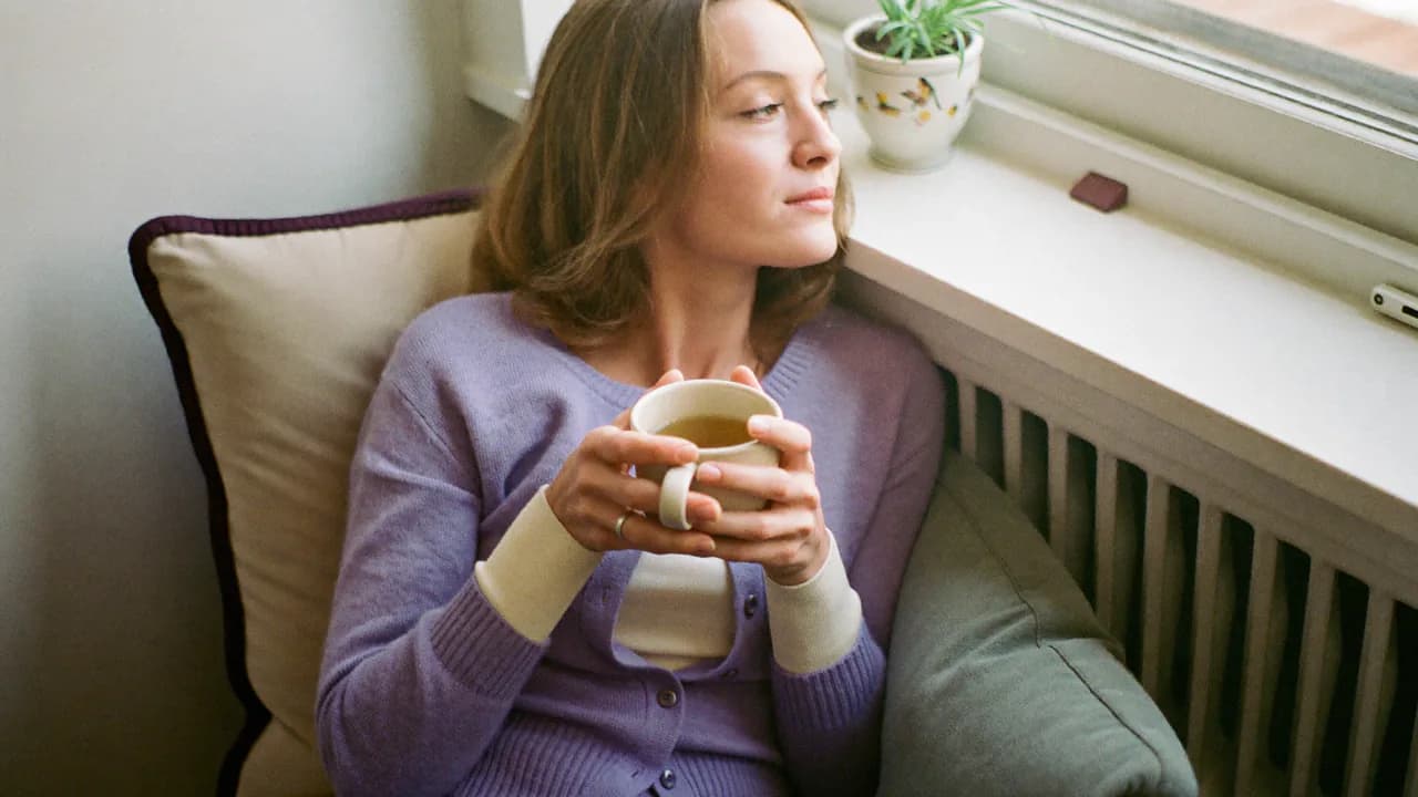 Une femme assise sur un banc de fenêtre chez elle, gilet lavande, une tasse de thé dans les mains, le regard tourné vers la fenêtre, l'air calme, dans la lumière du matin