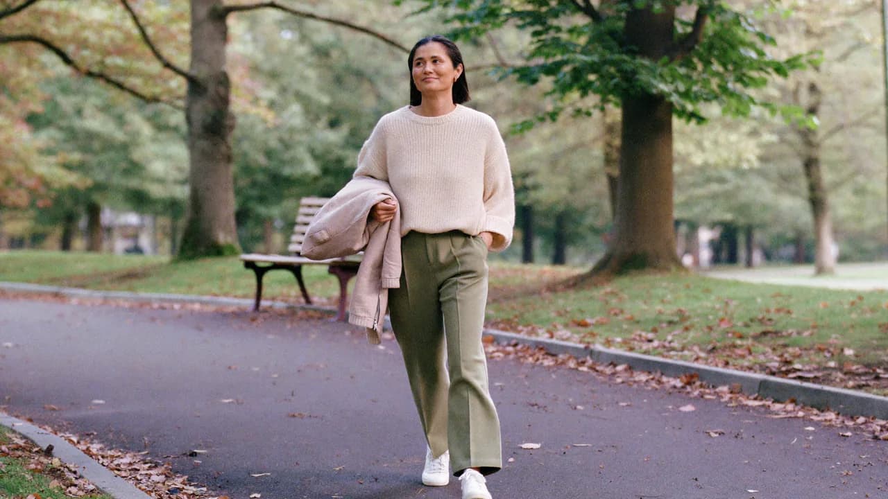 Une femme marche d'un pas tranquille dans une allée de parc bordée d'arbres, en pull crème et pantalon vert sauge, une veste sur le bras