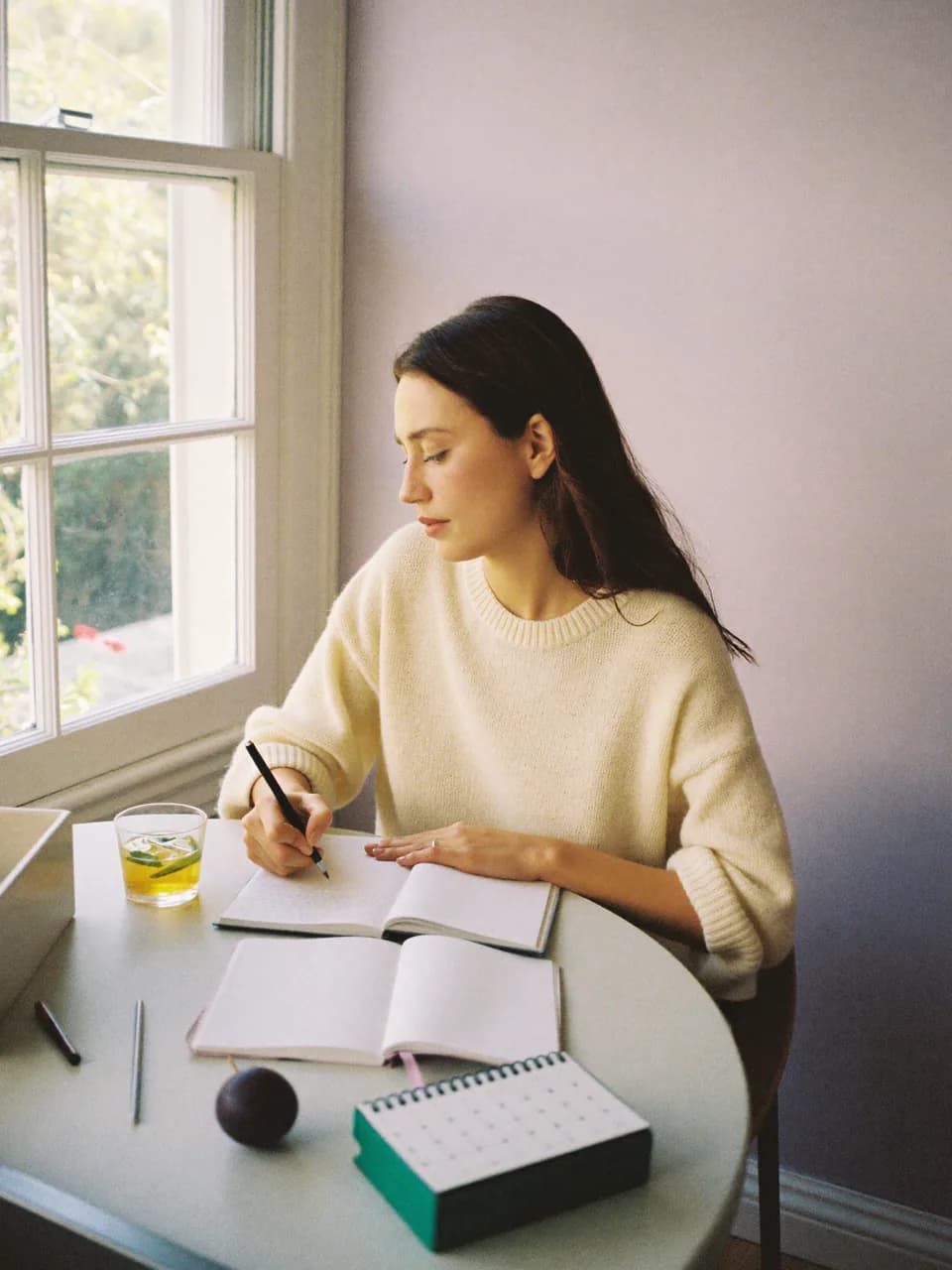 Une jeune femme écrit dans un carnet à une table de cuisine, une tisane et un calendrier à côté d'elle